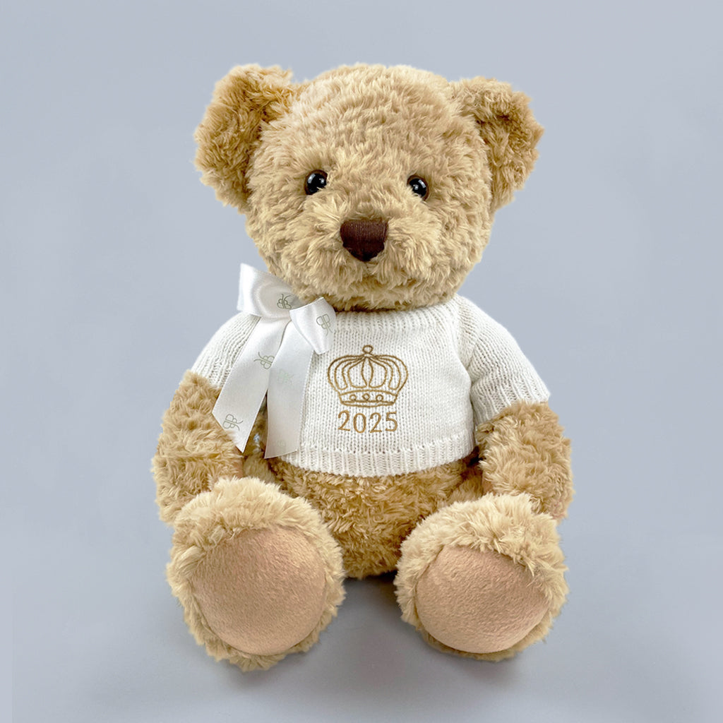 A traditional style seated teddy bear with a white knitted jumper embroidered with a crown or tiara design and the year 2025 in gold thread.