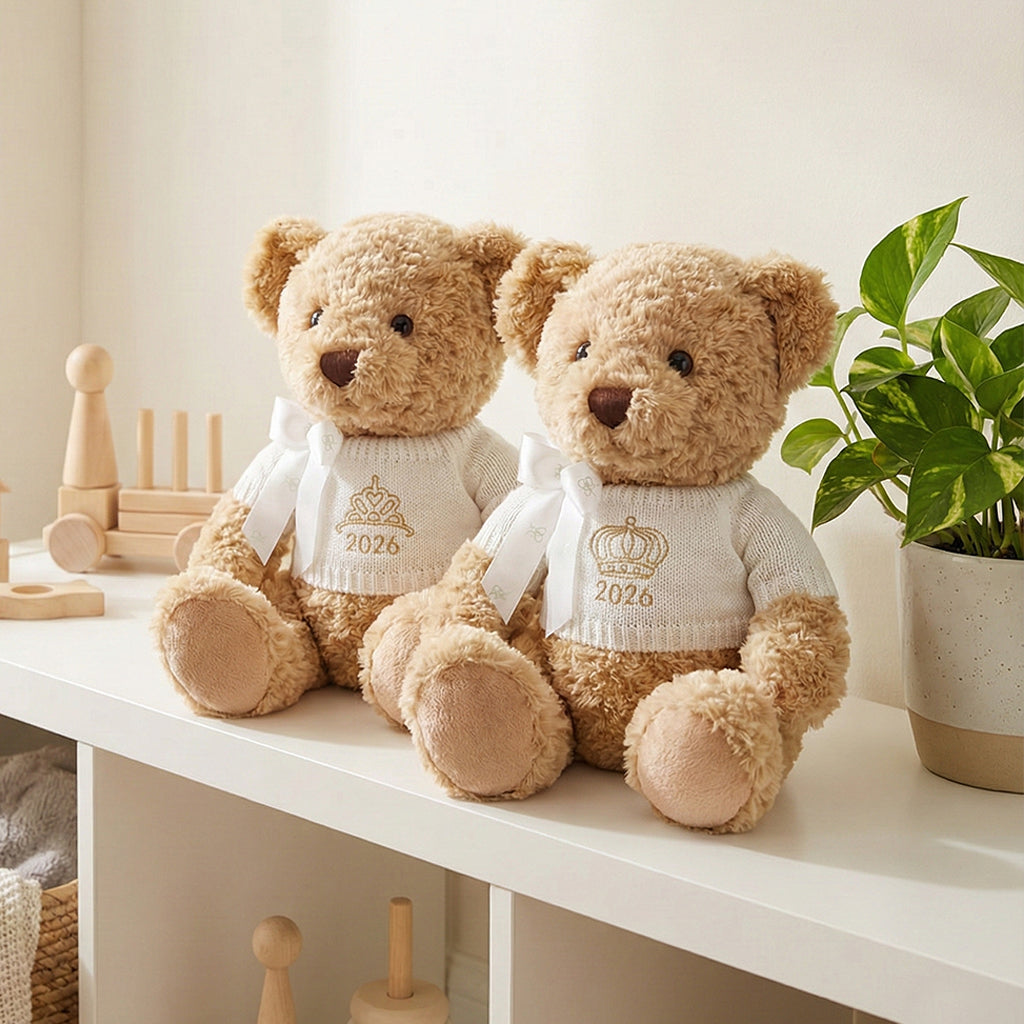 A traditional style seated teddy bear with a white knitted jumper embroidered with a crown or tiara design and the year 2026 in gold thread.
