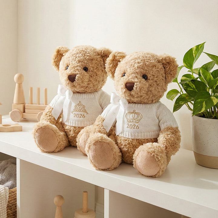 A traditional style seated teddy bear with a white knitted jumper embroidered with a crown or tiara design and the year 2026 in gold thread.