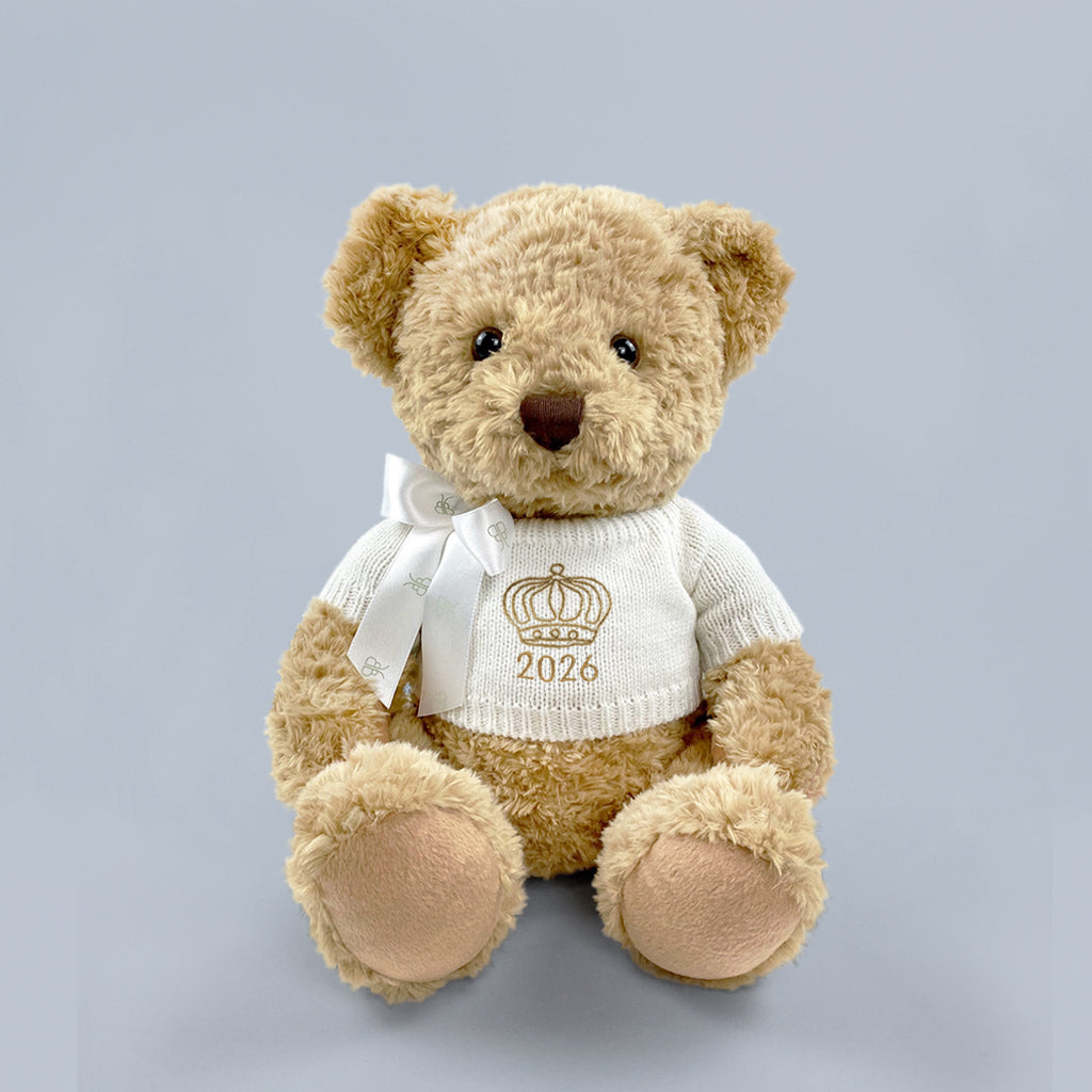A traditional style seated teddy bear with a white knitted jumper embroidered with a crown or tiara design and the year 2026 in gold thread.