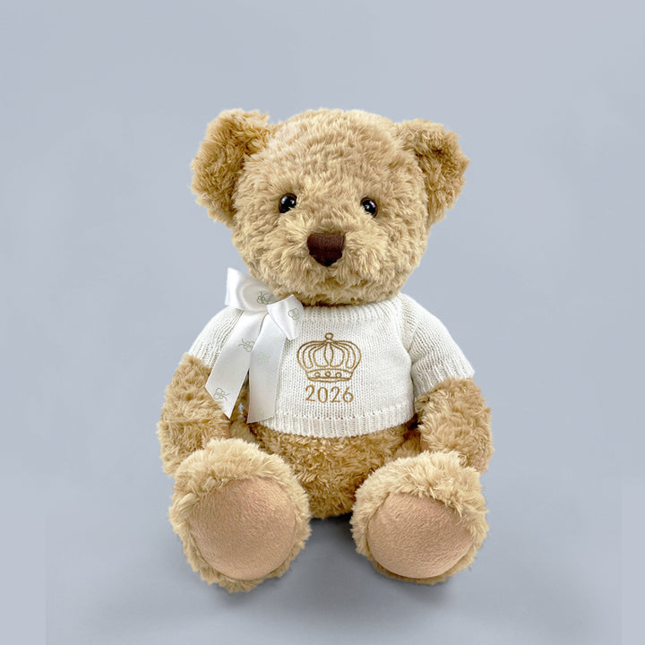 A traditional style seated teddy bear with a white knitted jumper embroidered with a crown or tiara design and the year 2026 in gold thread.