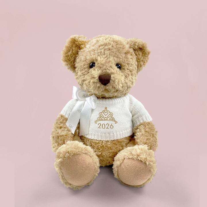 A traditional style seated teddy bear with a white knitted jumper embroidered with a crown or tiara design and the year 2026 in gold thread.