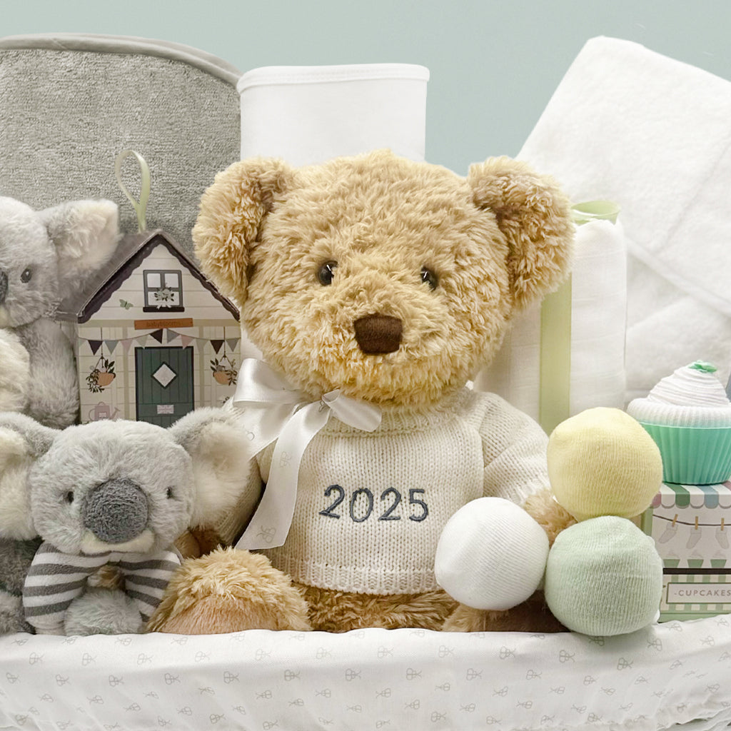Teddy bear wearing a knitted jumper with '2025' on it surrounded by baby gifts 