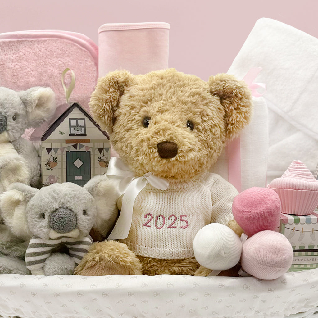 Teddy bear wearing a knitted jumper with '2025' on it, surrounded by soft toys and baby items.