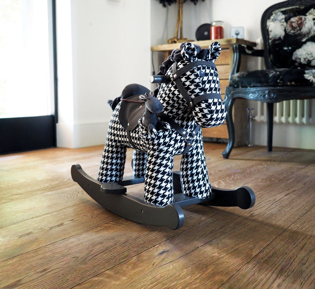 A black and white houndstooth patterned rocking horse with a leatherette saddle and bridle, sitting on a grey wooden frame with detachable footplates and easy-to-reach handles, with a smaller houndstooth-patterned horse toy attached.
